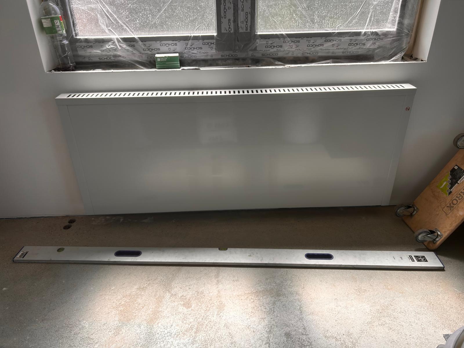 Nachtspeicher und Flächenspeicherheizung – der Unterschied in vier Punkten Weiße Flächenspeicherheizung, 162 cm breit und 8 cm tief, unter einem Fenster an der Wand montiert, davor eine 2-Meter-Wasserwaage als Größenvergleich