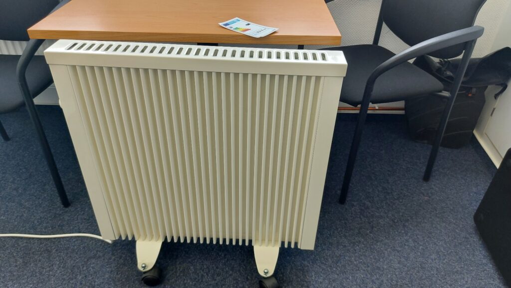 Mobile Flächenspeicherheizung auf Füßen mit Rollen neben einem Schreibtisch im Büro