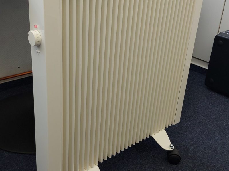 Weiße Flächenspeicherheizung auf Füßen mit Rollen im Büro, Temperaturregler an der Seite